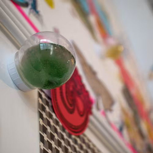 A close up photo of a bubble-shaped glass jar filled with murky green liquid, which is attached to a white wall. The wall is blurry, and covered in colourful paper cutouts.