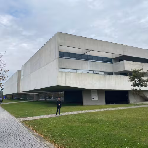Exterior of a large concrete brutalist building, with blue sky, green lawn, and tree lined sidewalk.