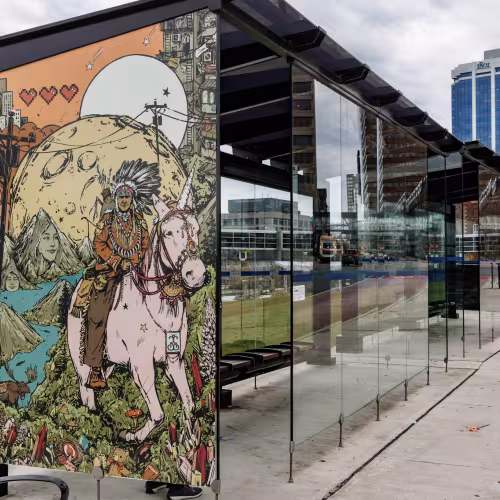Image is of the transit terminal shelter with artwork from 2025 Interchange artist Zeta Paul, titled &ldquo;Msɨt No&rsquo;kmaq (All My Relations): Everything is Smiling&rdquo;. There is a colourful illustration with an Indigenous man riding a pale pink unicorn in the foreground, wearing a war bonnet, full regalia, and a bow and arrows on his back. He is riding towards the viewer over a hill of green grass covered with mushrooms, teddy bears, lupins, and pink crystals. The unicorn wears a bejewelled harness and around its&hellip;